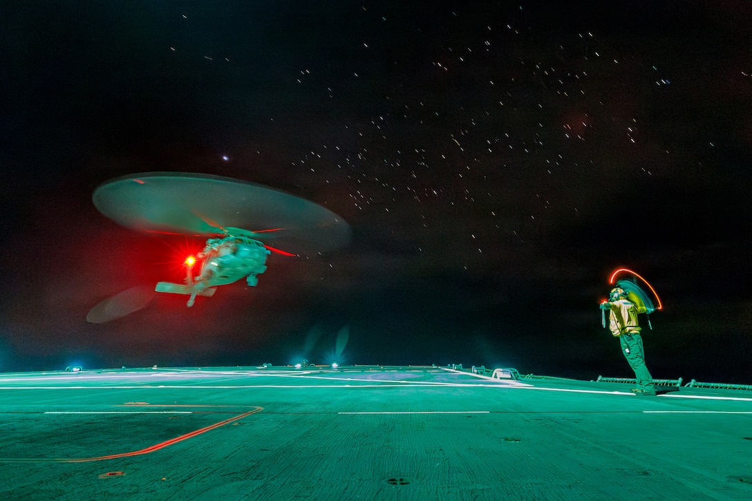 The rotor blades of a helicopter look like a flying saucer against a black starry sky as it comes in for a landing on the deck of a ship. A sailor signals to a pilot with red lights.