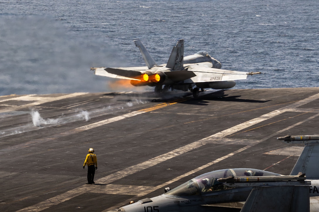A fighter jet with smoke behind it is on the edge of a runway, with water in the background.