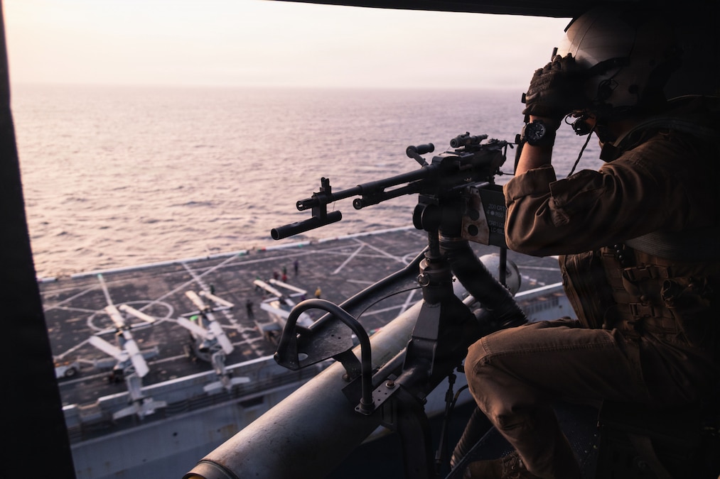 A Marine looks out of an open door of a helicopter, with a weapon nearby and the flight deck of a ship and water below the Marine.