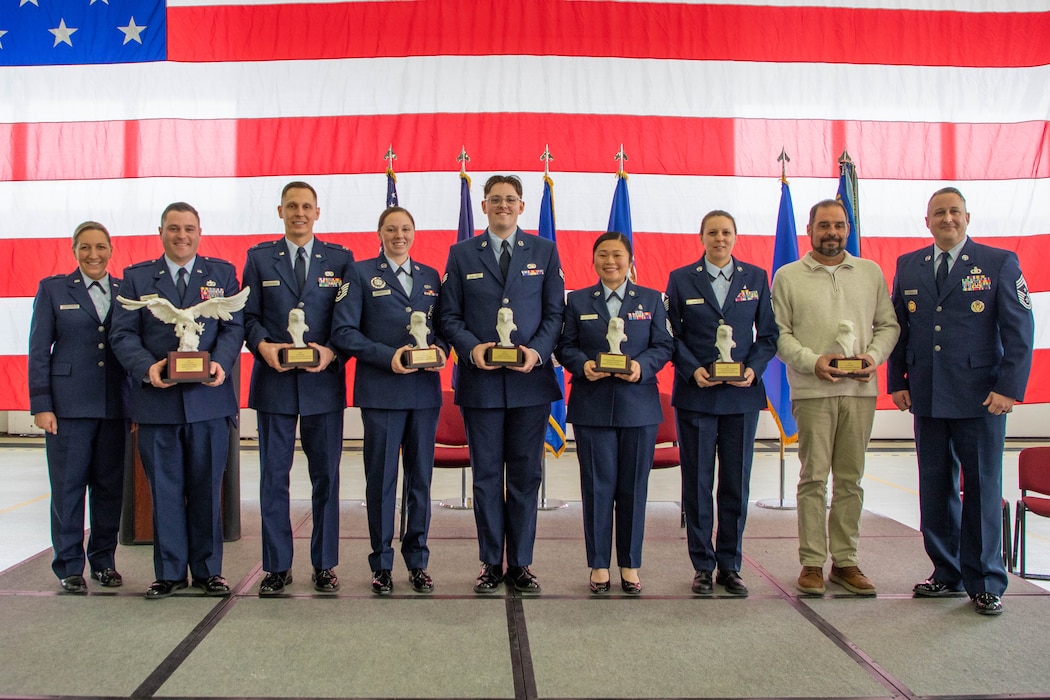 Eight uniformed Airmen and one civilian stand in a line, holding trophies