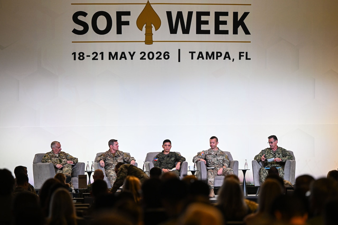 Five men wearing camouflage military uniforms sit indoors in chairs on a stage facing a crowd of people with their backs to the camera. A sign above the men on the wall reads, "SOF Week, May 18-21, 2026, Tampa, Florida."