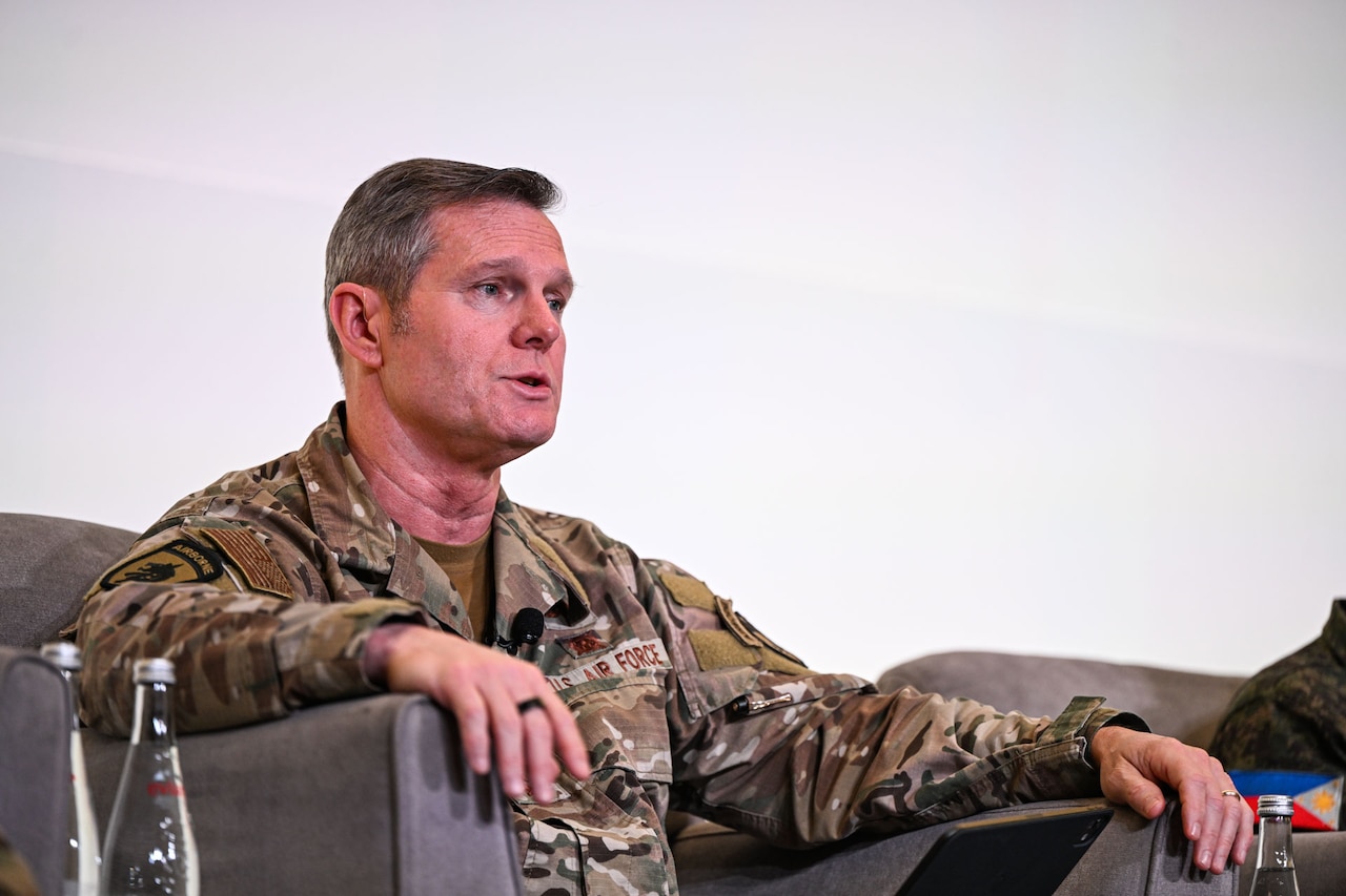 A man wearing a camouflage military uniform speaks on stage while sitting in a chair.
