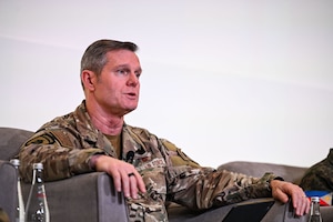 A man wearing a camouflage military uniform speaks on stage while sitting in a chair.