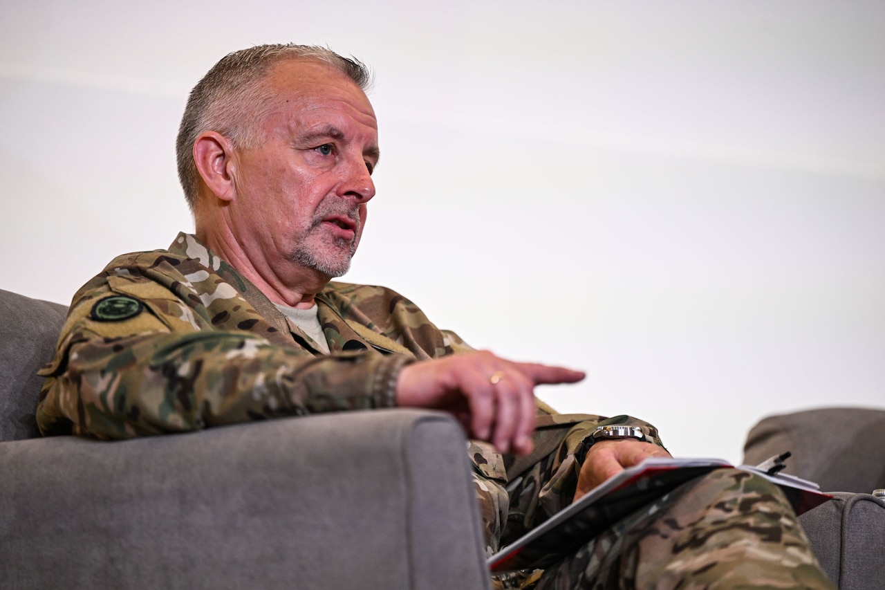 A man wearing a camouflage military uniform speaks on stage while sitting in a chair.