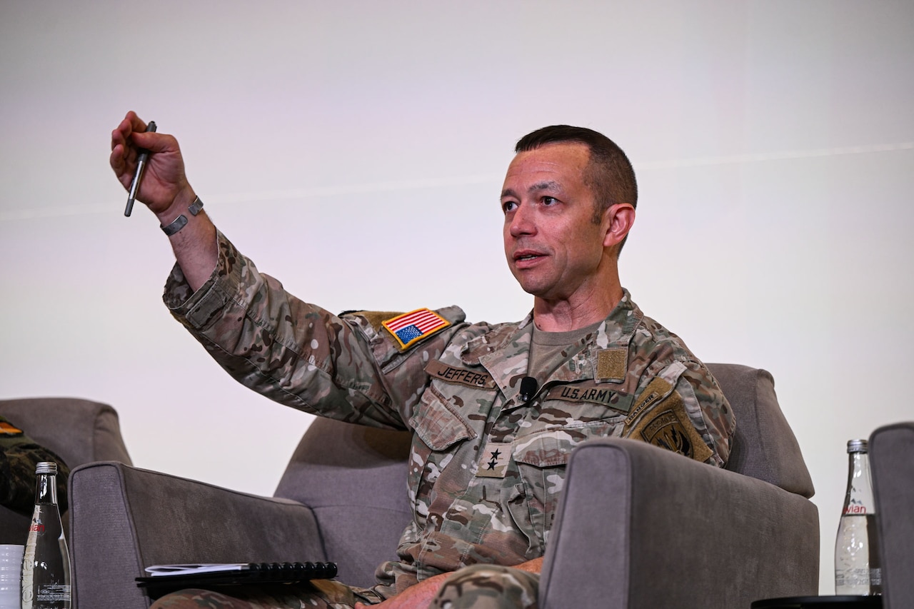 A man wearing a camouflage military uniform speaks on stage while sitting in a chair.