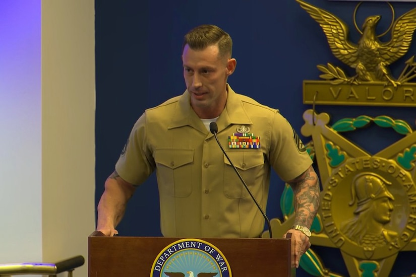 A Marine in a short-sleeved military dress shirt speaks into a microphone while standing behind a lectern indoors.
