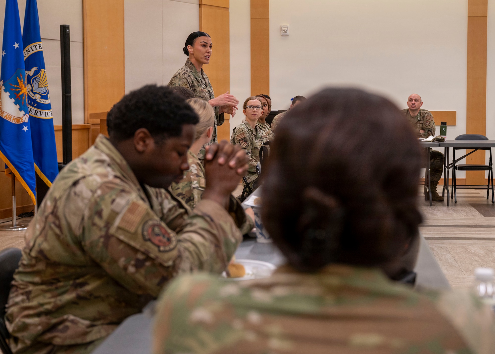 Military members are seated at a "U" shaped table listening to a woman at the head of the table. She's standing and gesturing with her hands in front of her.