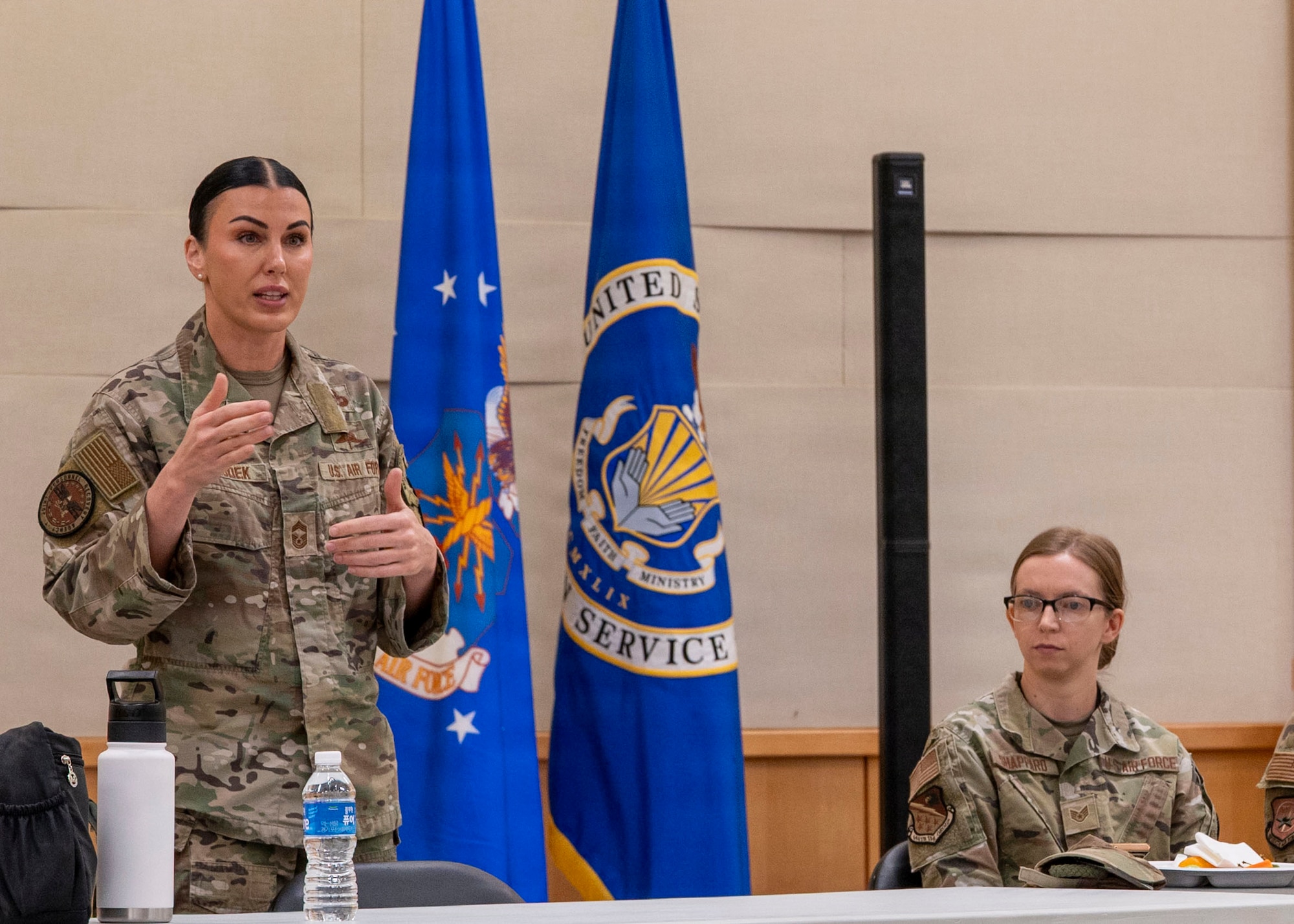 Two women in OCP camouflage uniforms are at a table. One is standing and gesturing with her hands. The other is on her left listening to what she's saying. There are flags and a standing speaker in the background.