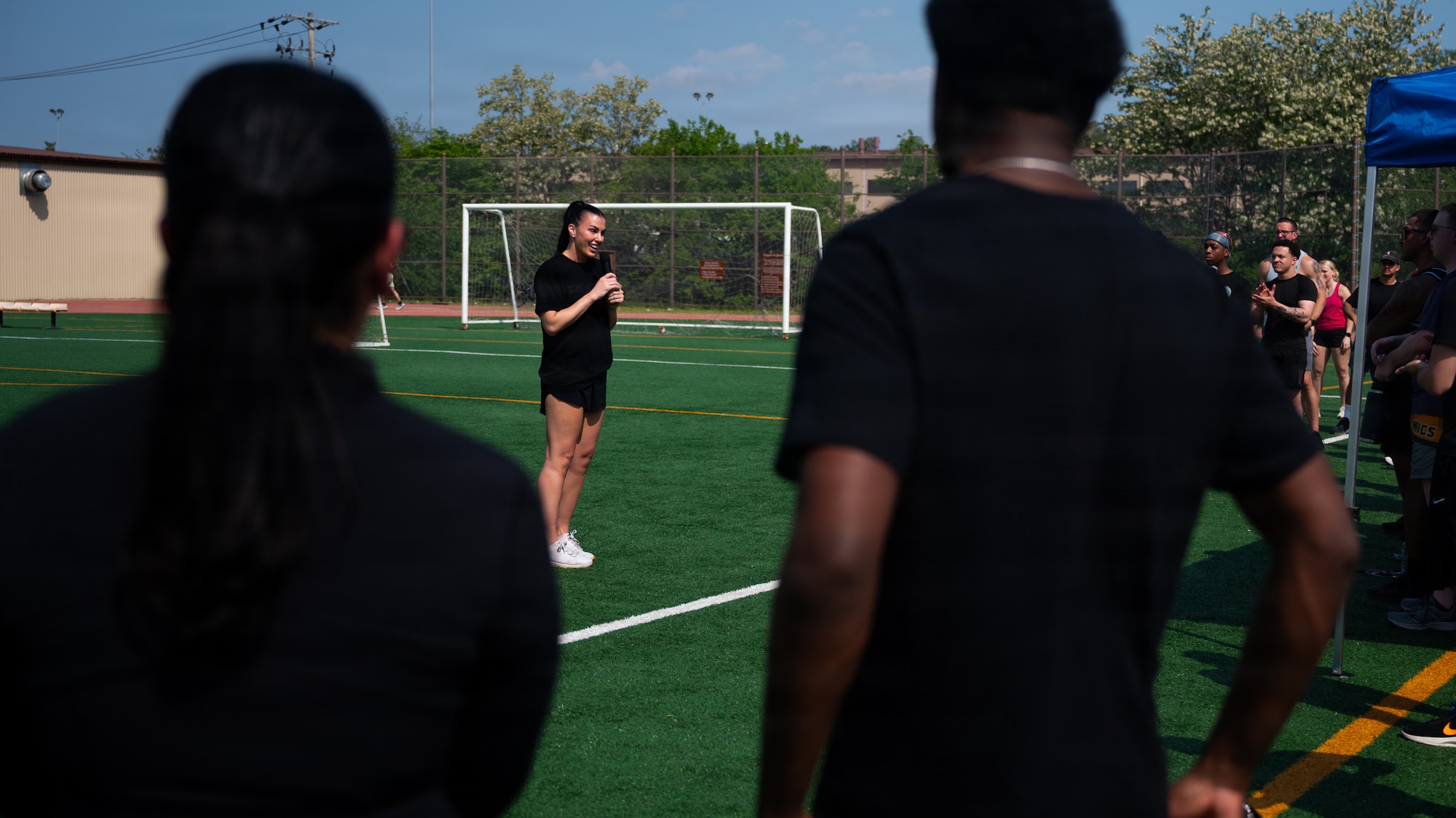 Woman in athletic clothing stands on a green soccer field with a mic in her hand. She's talking to a crowd of people.
