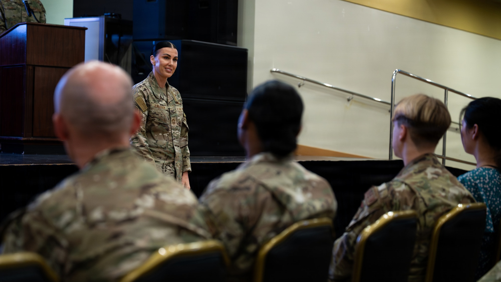 Woman in OCP camouflage uniform stands smiling in front of people in chairs.