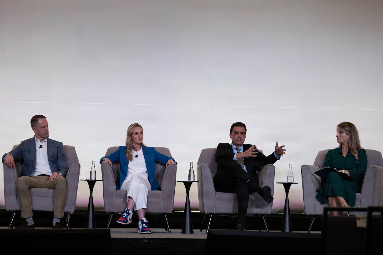 Four civilians  in business attire sit in chairs on a stage while one talks and gestures.