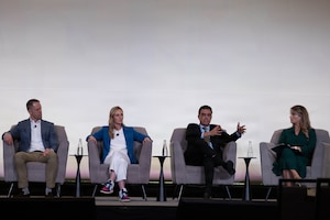 Four civilians  in business attire sit in chairs on a stage while one talks and gestures.