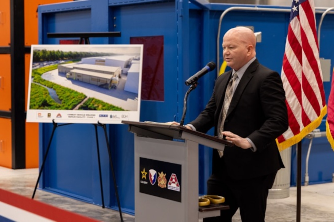 Picture of a man standing at a podium speaking with a picture of a rehab is in the background.