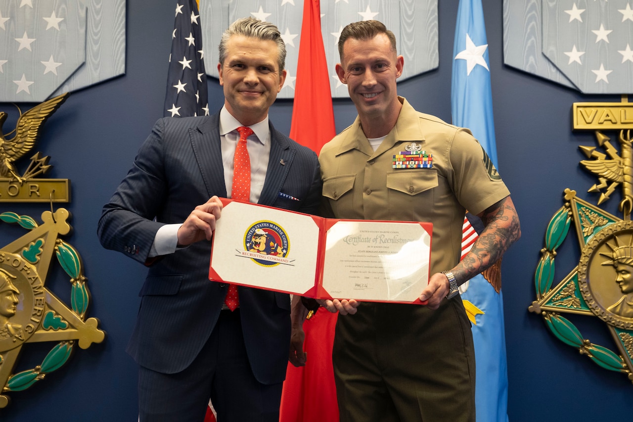 Secretary of War Pete Hegseth and a Marine jointly hold up a certificate and smile for a photo.