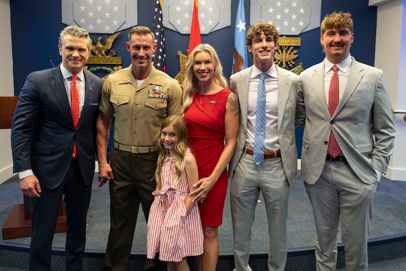 Five people in business attire and a man in a formal military uniform pose for a photo. Behind them are assorted flags and symbols on a wall.