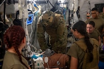 Three military medical professionals are standing over a patient on a gurney. A chaplain has his arms on the patient's chest and is performing CPR. There is medical equipment connected to the patient.