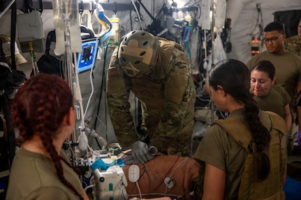 Three military medical professionals are standing over a patient on a gurney. A chaplain has his arms on the patient's chest and is performing CPR. There is medical equipment connected to the patient.