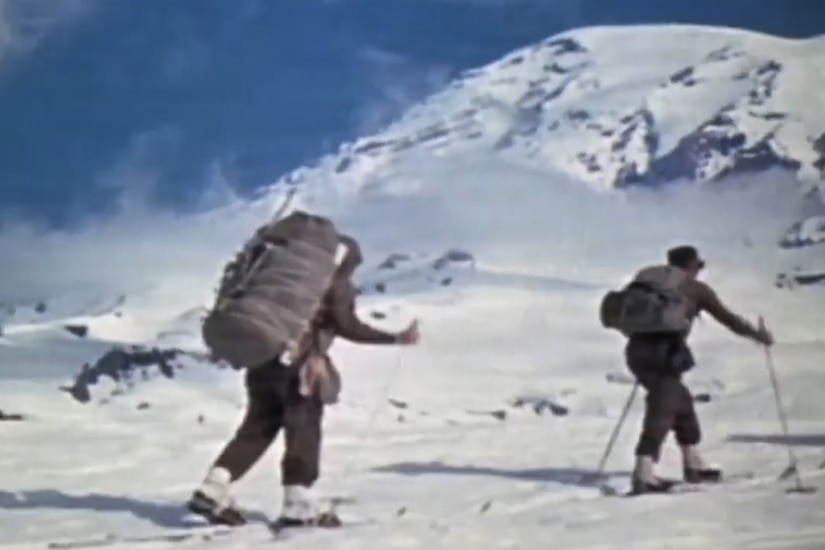 Two men wearing historical military winter gear walk through a snow-covered mountainous area.