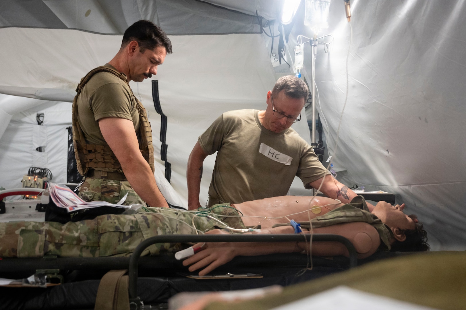 Two chaplains stand over a patient in a tent and they are praying over the patient and checking vital signs.