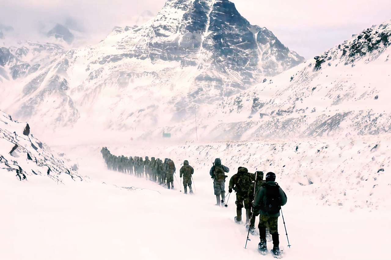 A long line of people dressed in military winter gear walk through a snow-covered mountainous area.