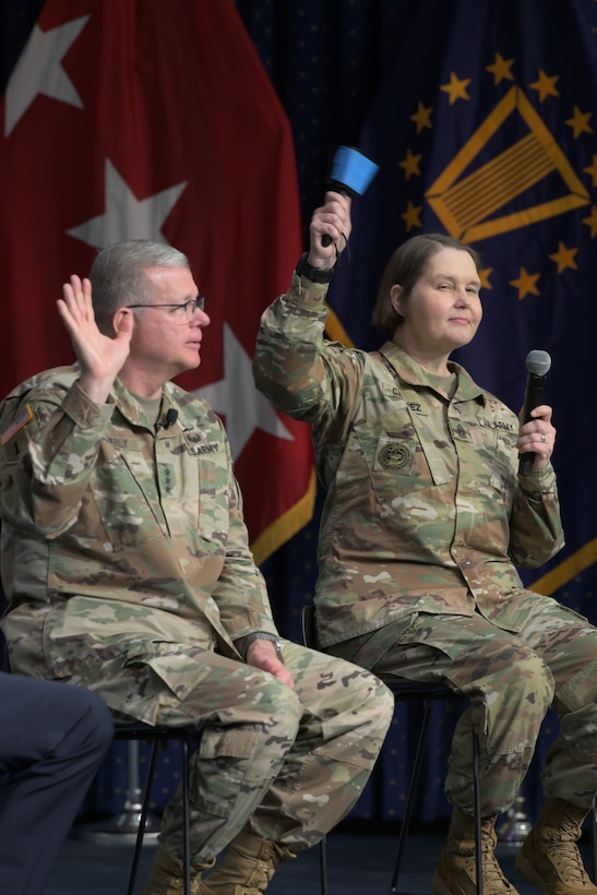 A man and a woman, both wearing Army uniforms, sit on stools on a stage.