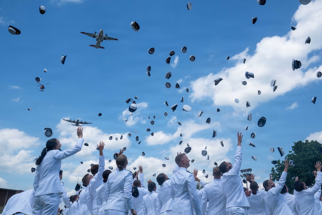 Dozens of Coast Guardsmen in ceremonial dress toss their graduation caps into the air as aircraft fly above in a blue sky.