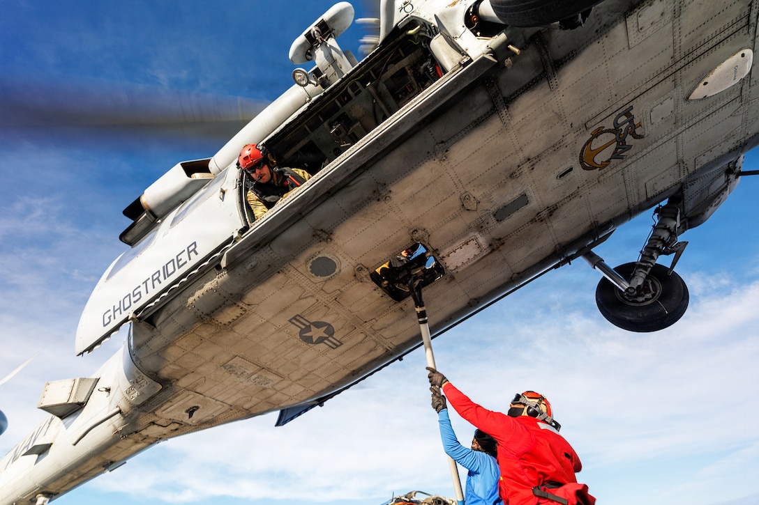 A sailor looks out the open doorway of an airborne helicopter as two fellow sailors attach a hook to it.