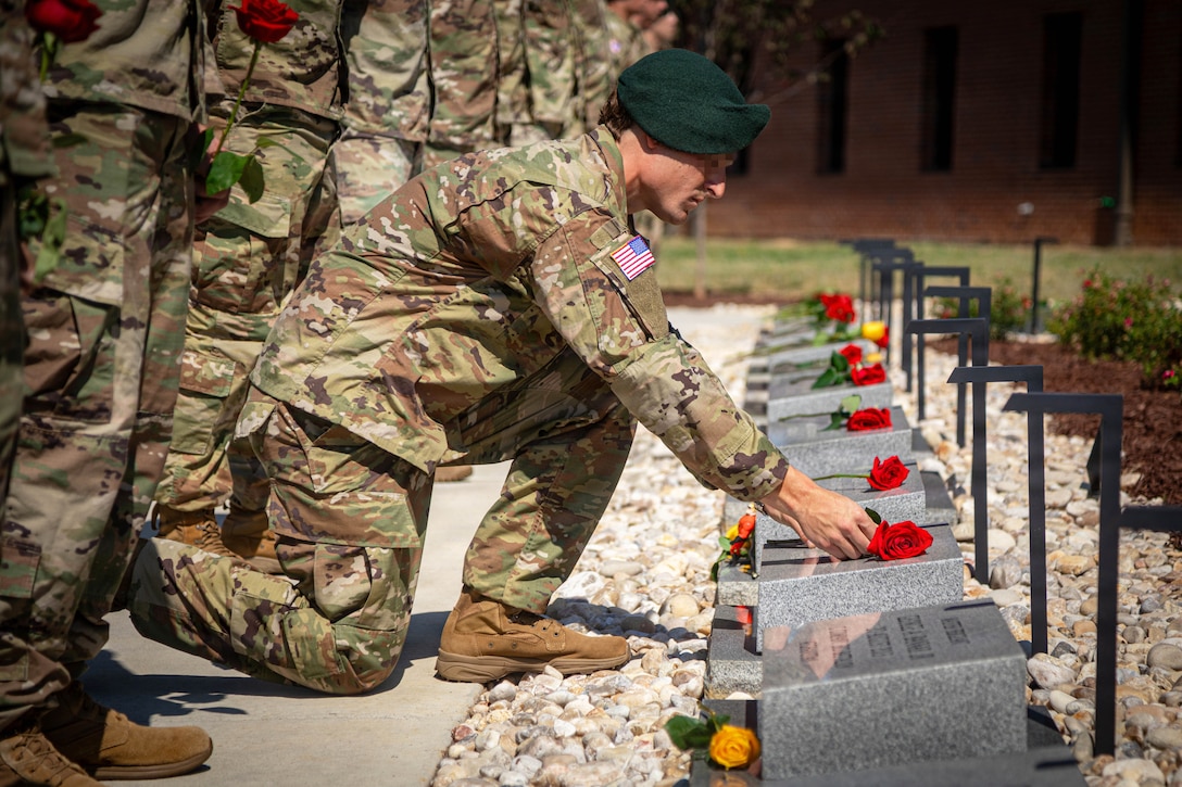 A soldier kneels next to fellow soldiers while placing a rose on a memorial stone during the day.