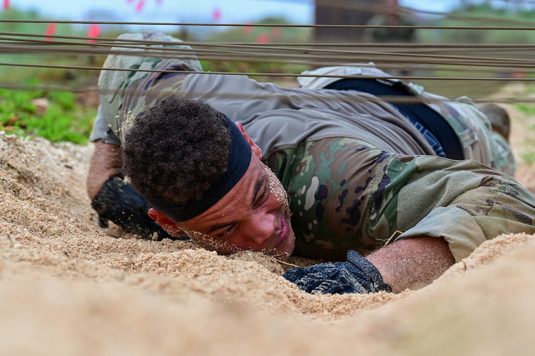 A soldier low-crawls under wire on sand. His face is turned to the side, and sand is covering both sides of it.
