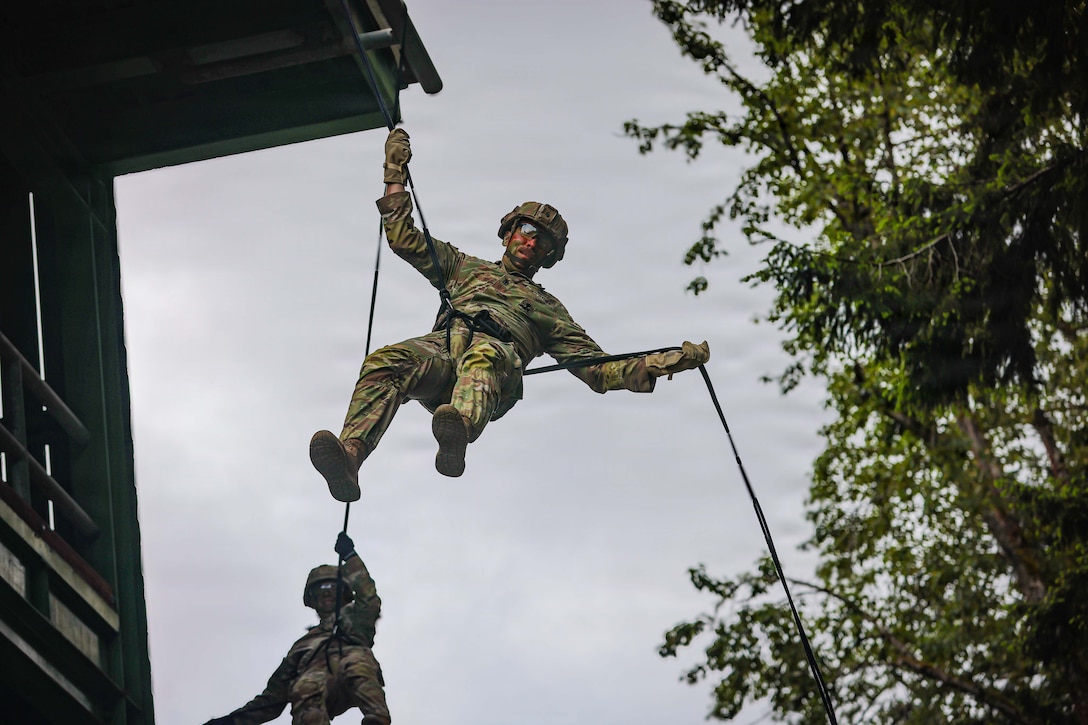 Two soldiers in seat harnesses hang from and hold onto a rope that is connecting them by the side of a building, with trees to their other side and a gray sky in the background.