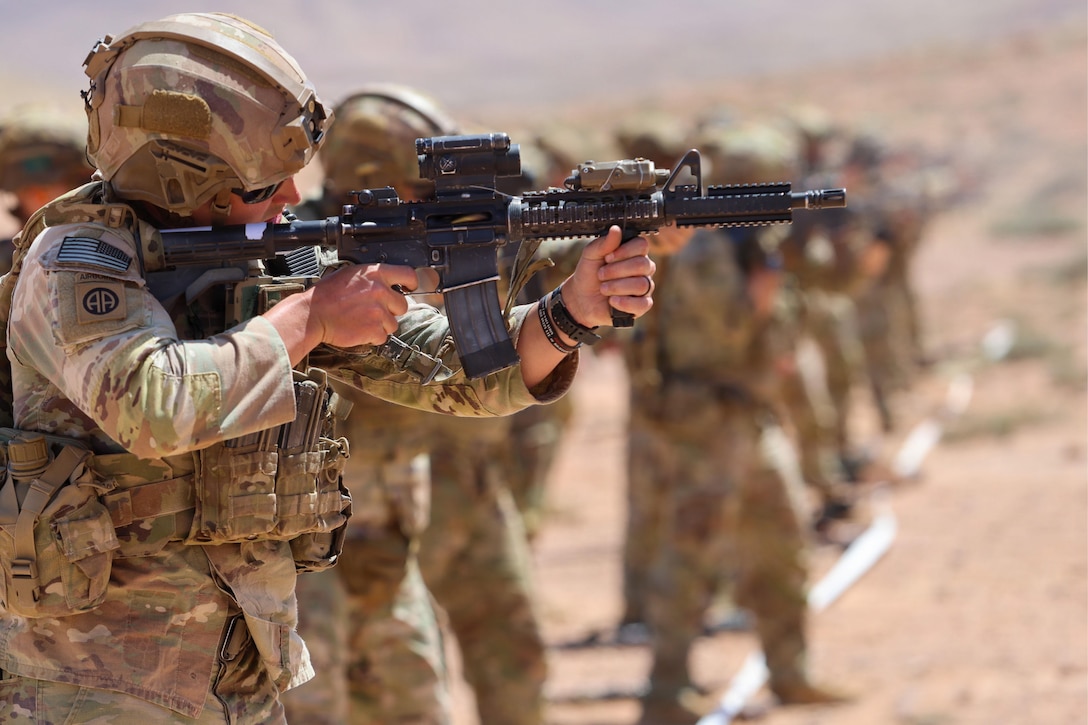 Soldiers stand in a row on dirt holding machine guns as if about to fire at targets. One soldier is in focus.
