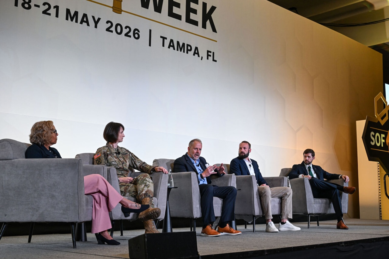 Four people wearing business attire and one in a camouflage military uniform sit in chairs on a stage. The man in the middle is talking and gesturing.