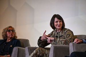 A woman wearing a camouflage military uniform sits in a chair while talking and gesturing as a woman in business attire listens.