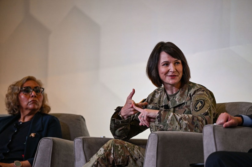 A woman wearing a camouflage military uniform sits in a chair while talking and gesturing as a woman in business attire listens.