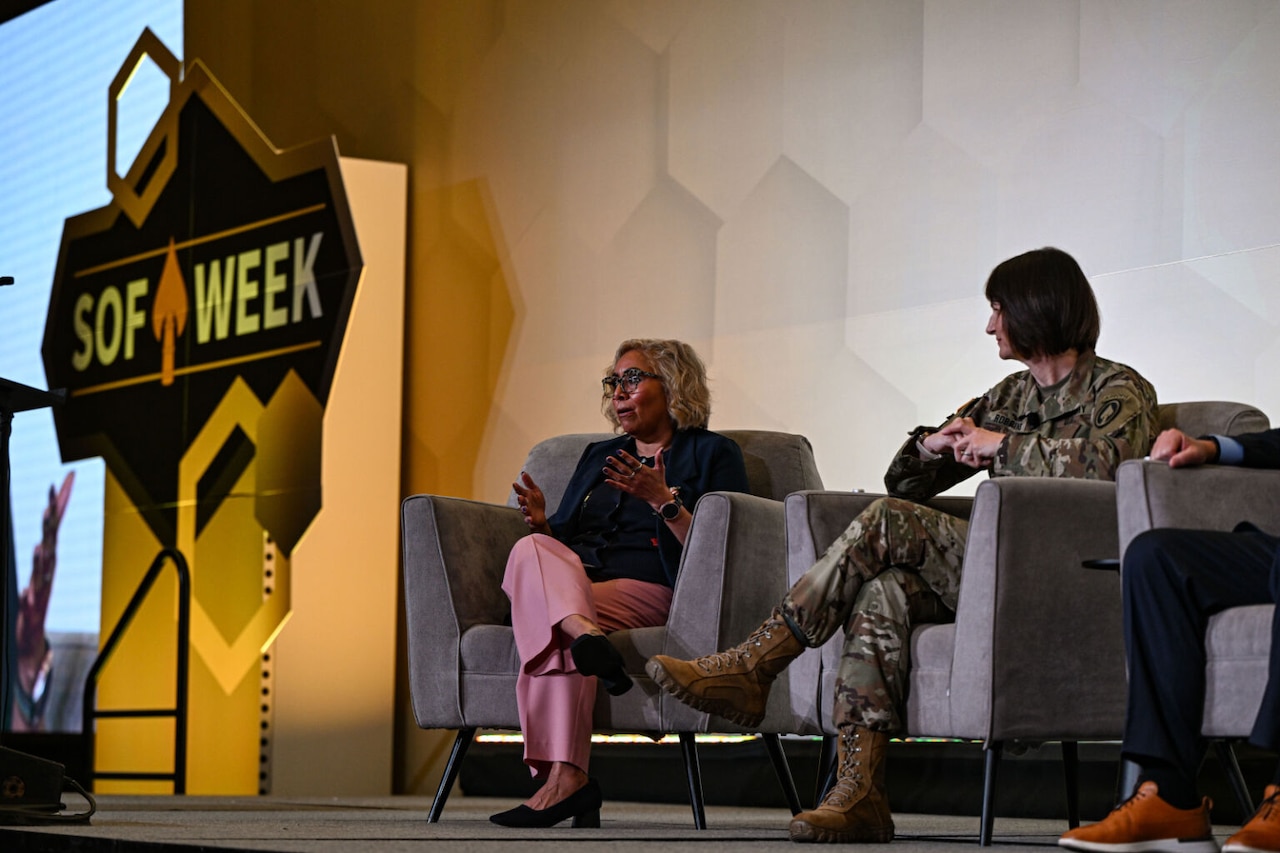 Two women, one wearing a camouflage military uniform and one in business attire, sit in chairs on a stage next to a sign that reads, “SOF Week.” The woman on the left speaks and gestures to an audience off camera.