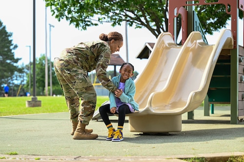 a woman laughs with a child at the bottom of a playground slide.
