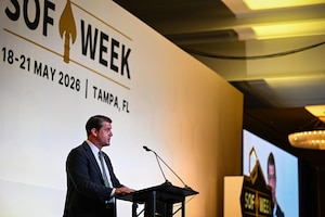 A man in business attire stands behind a lectern and speaks into a microphone. Behind him, a sign on the wall reads, "SOF Week, 18-21 May 2026, Tampa, FL."