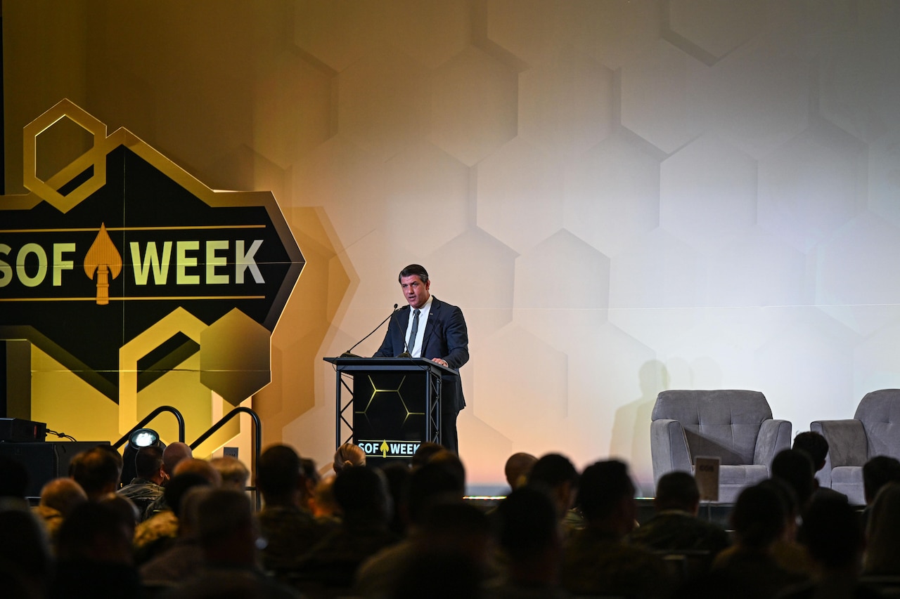 A man in business attire stands behind a lectern and speaks into a microphone. In front of him is a seated audience with their backs to the camera.