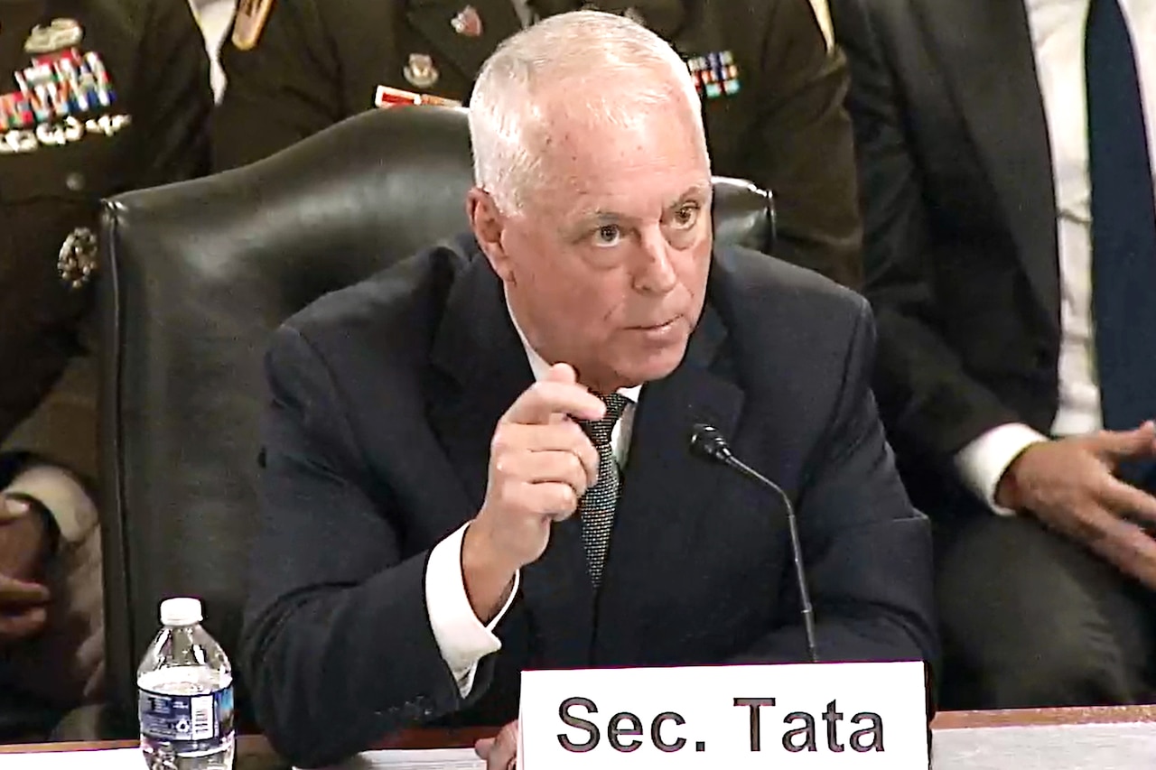 A man wearing business attire sits at a table and speaks into a microphone; in front of him is a place card that reads, "Sec. Tata."