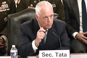 A man wearing business attire sits at a table and speaks into a microphone; in front of him is a place card that reads, "Sec. Tata."
