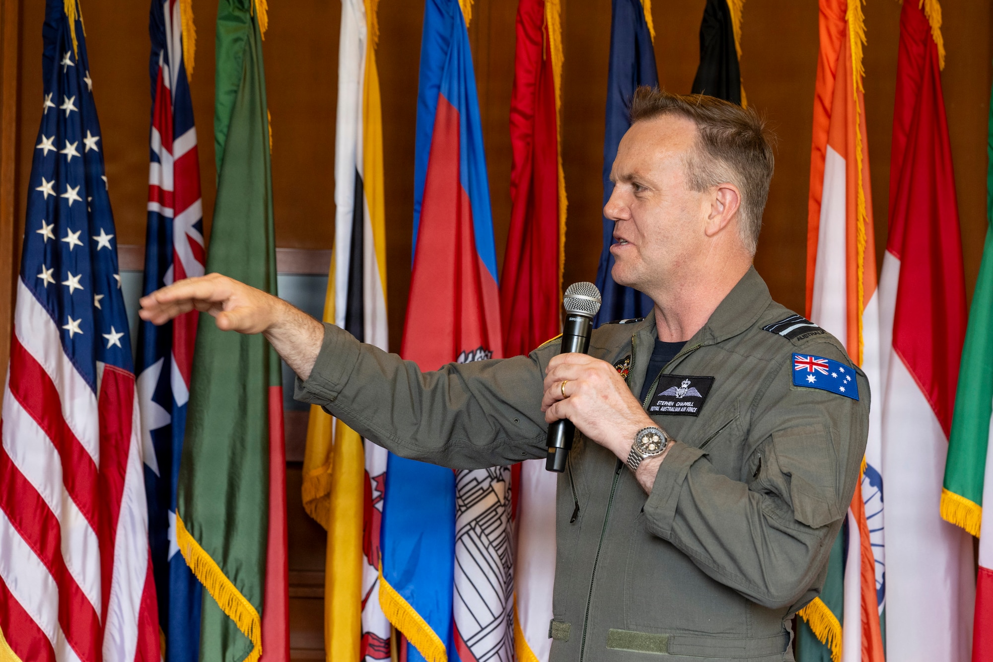 Royal Australian Air Force Air Marshal Stephen Chappell, Chief of Air Force, gestures with his hand as he speaks into a microphone. He is standing in front of a colorful backdrop of numerous international flags, including a prominent American flag.
