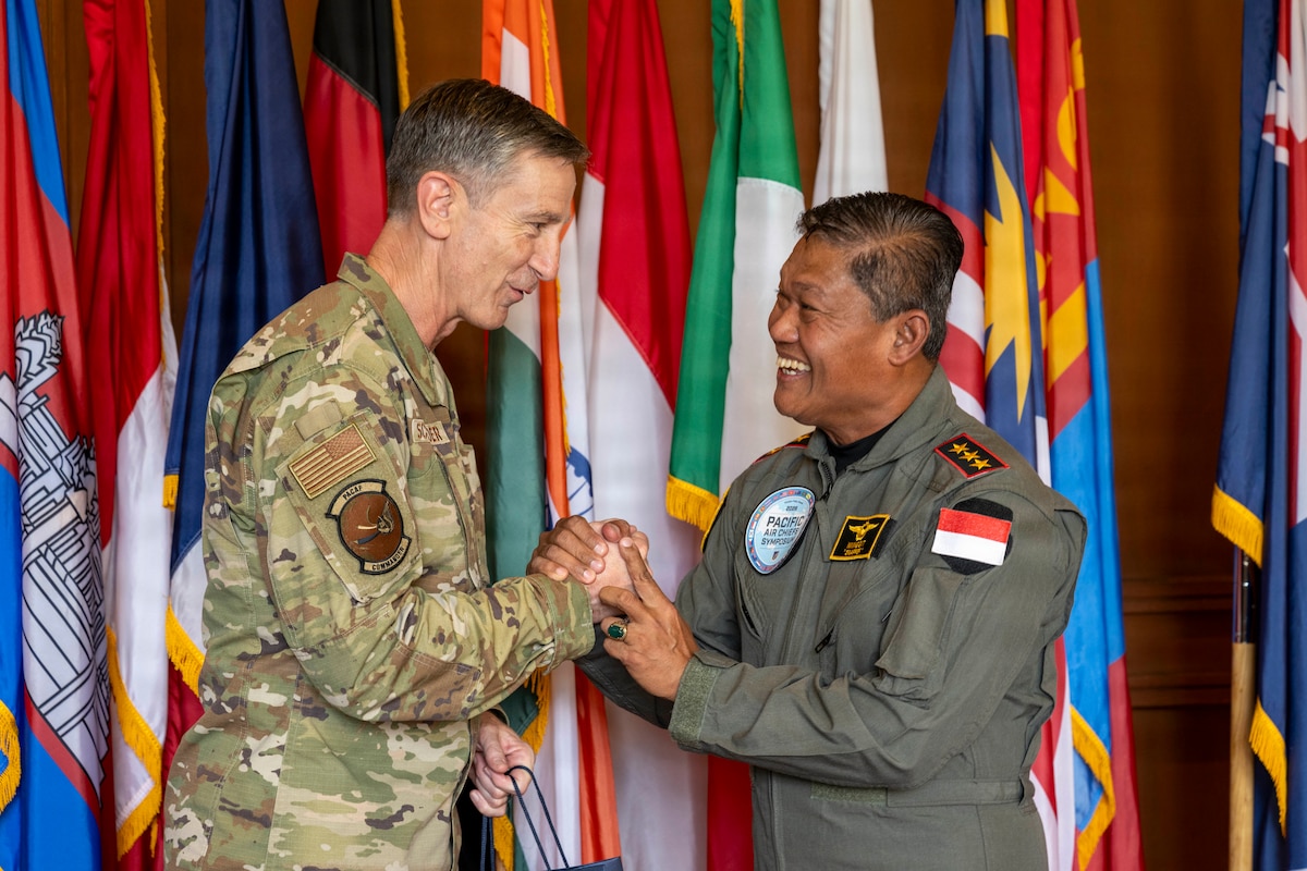 U.S. Air Force Gen. Kevin Schneider, Pacific Air Forces commander, smiles as he clasps hands with Indonesian Air Force Air Marshal Mingget Tribowo. The two leaders are standing in front of a vibrant backdrop of international flags.