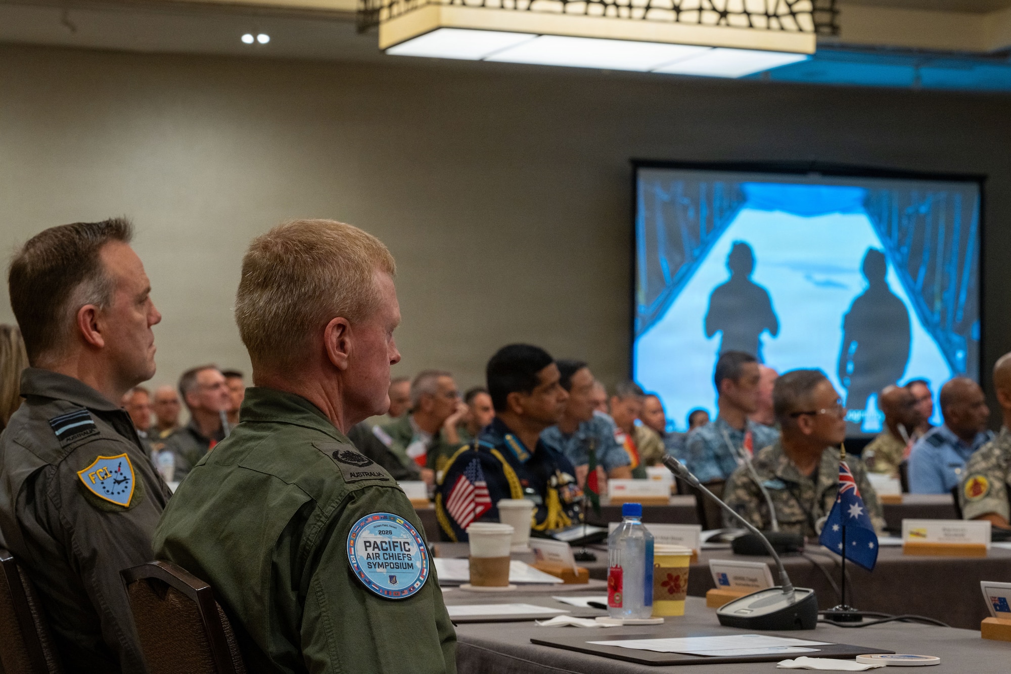 From a viewpoint within the audience, multinational air force leaders watch a presentation on a large screen during a symposium. A service member in the foreground has a prominent 'Pacific Air Chiefs Symposium' patch on their sleeve, and small international flags are visible on the conference tables.