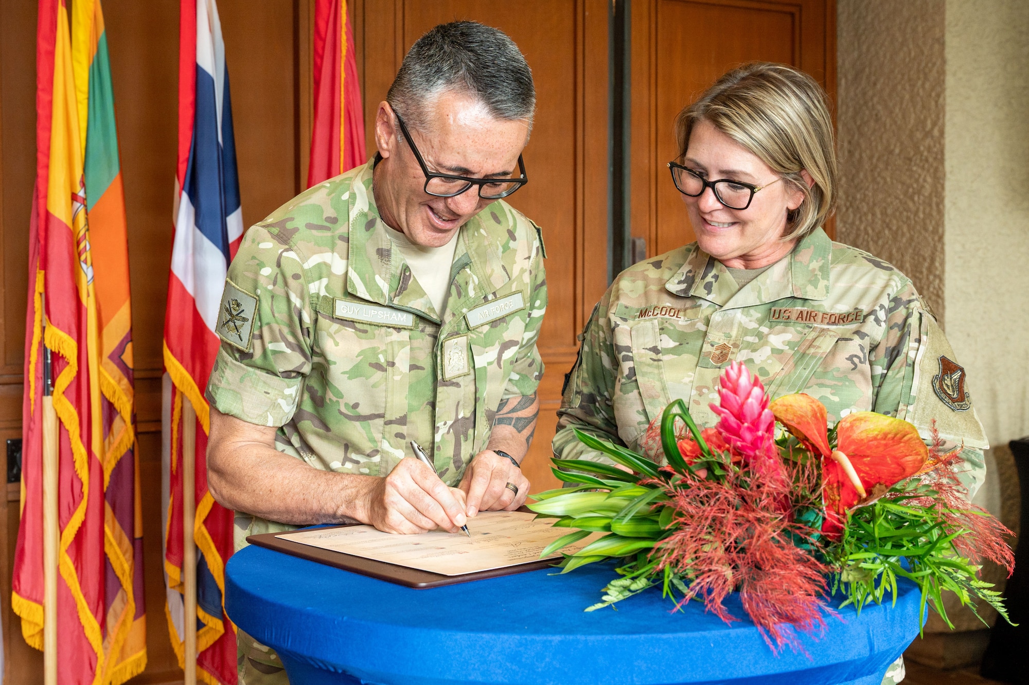 U.S. Air Force Command Chief Master Sgt. Katie McCool smiles as she looks on while Royal New Zealand Warrant Officer of the Air Force Dwayne Lipsham signs a document on a table. The two are in uniform, standing behind a table with a large tropical flower arrangement and a backdrop of international flags.