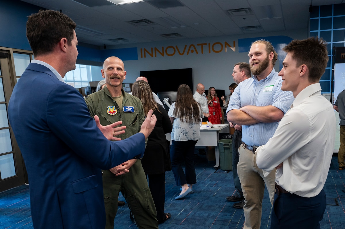 Males stand in group and chat. one is wearing an Air Force flight suit.