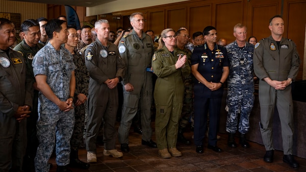 A large, diverse group of multinational military leaders stands together while listening to closing remarks. The attendees are wearing a wide variety of uniforms, including flight suits and different camouflage patterns, with one woman in the center of the group clapping.