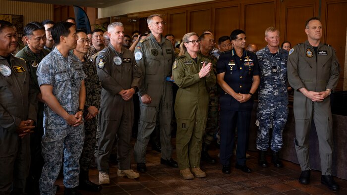 A large, diverse group of multinational military leaders stands together while listening to closing remarks. The attendees are wearing a wide variety of uniforms, including flight suits and different camouflage patterns, with one woman in the center of the group clapping.