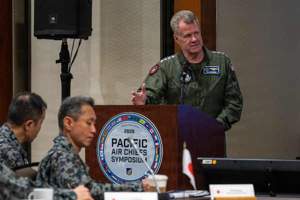 "U.S. Navy Adm. Samuel J. Paparo, in a flight suit, gestures while speaking from behind a wooden podium. The podium features a large, circular seal for the 'Pacific Air Chiefs Symposium,' ringed with international flags. In the foreground, attendees from other nations are seated at a conference table, listening.