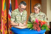 U.S. Air Force Command Chief Master Sgt. Katie McCool smiles as she looks on while Royal New Zealand Warrant Officer of the Air Force Dwayne Lipsham signs a document on a table. The two are in uniform, standing behind a table with a large tropical flower arrangement and a backdrop of international flags.