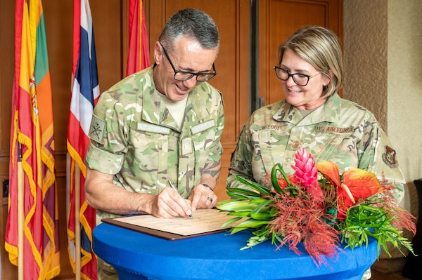 U.S. Air Force Command Chief Master Sgt. Katie McCool smiles as she looks on while Royal New Zealand Warrant Officer of the Air Force Dwayne Lipsham signs a document on a table. The two are in uniform, standing behind a table with a large tropical flower arrangement and a backdrop of international flags.